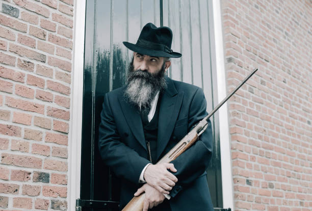 istock Man with hat and long beard standing with rifle against window shutters. 686205178