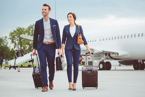 Full length shot of two young business people walking in front of airplane with luggage. People in business trip.