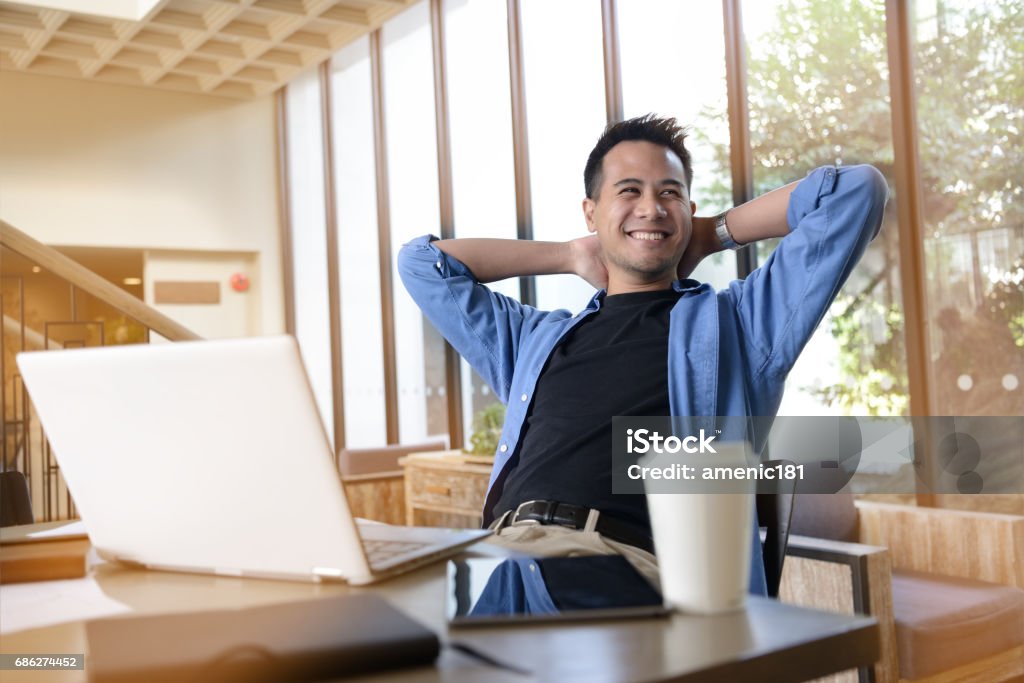 Glücklicher junger Geschäftsmann arbeitet am Laptop, während er an seinem Arbeitsplatz im Büro sitzt - Lizenzfrei Asiatischer und Indischer Abstammung Stock-Foto