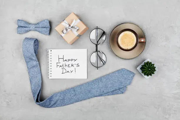 Happy Fathers Day background with morning coffee mug, gift, glasses, necktie and bowtie on stone table top view in flat lay style.