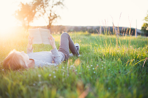 Young girl reading a book in the grass while sun setting down. Summer feeling.