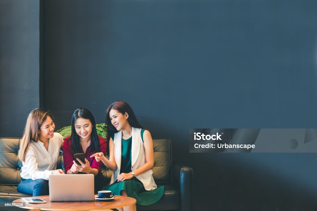 Tres hermosas asiáticas chicas utilizando smartphone y ordenador portátil, charlando en el sofá en la cafetería con espacio de copia, estilo de vida moderno con tecnología gadget o mujer trabajadora en concepto de negocios informal - Foto de stock de Amistad libre de derechos