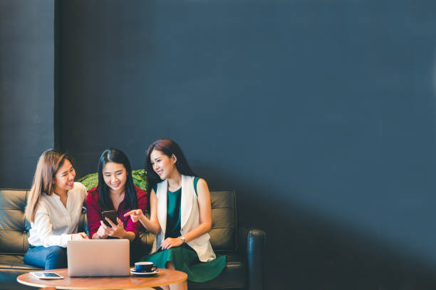 Three beautiful Asian girls using smartphone and laptop, chatting on sofa at cafe with copy space, modern lifestyle with gadget technology or working woman on casual business concept