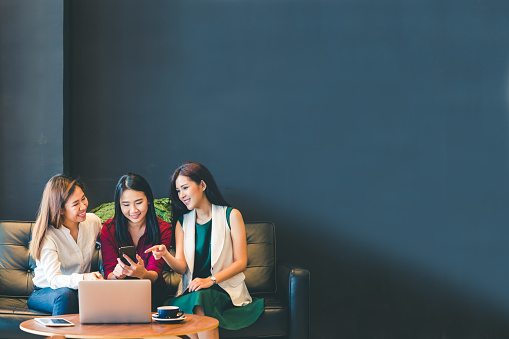 Three beautiful Asian girls using smartphone and laptop, chatting on sofa at cafe with copy space, modern lifestyle with gadget technology or working woman on casual business concept