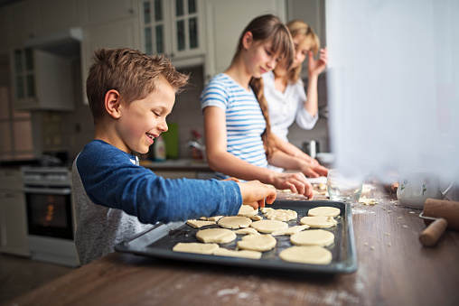 Family is having fun making cookies. Mother and daughter are cutting out cookies out of rolled out dough and the little boy is laying out the cookies on the baking tray. Kids are aged 11 and 7.\n