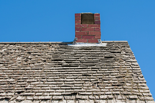 A very old wood shingle roof in poor repair. A chimney is on the top of the roof. Moss and lichens are growing from the roof. The sky above is blue.