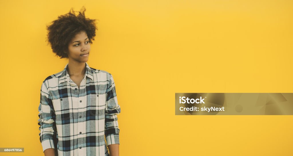 Black girl with copy space on yellow background Teenage black model in casual outfit pensively looking aside while standing in front of yellow background, serious young Brazilian girl in plaid shirt with copy space for text, logo or advertising Teenager Stock Photo