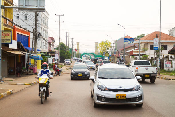 Morning traffic scene in Vientiane. In strete cars and a motorcycle driver are passing scene. In background are buildings.