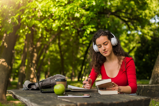 Serious Young woman, student  reading a book  with happy face listening music with white headphones, sitting by wooden table with stack of colorful hardback books,  her notes, laptop, backpack, tablet, green apple on blurred nature landscape backdrop during bright sunny day . Copy space for text