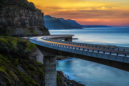 Sunset over the Sea cliff bridge along Australian Pacific ocean coast with lights of passing cars near Sydney, Australia. Long exposure.