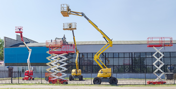 Several different wheeled scissor lifts and wheeled articulated lifts with telescoping boom and basket on an asphalt ground against the sky and an industrial building