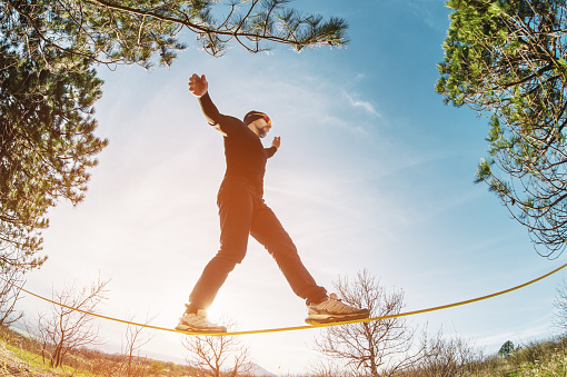A man, aged with a beard and wearing sunglasses, balances on a slackline in the open air between two trees at sunset on background blue sky