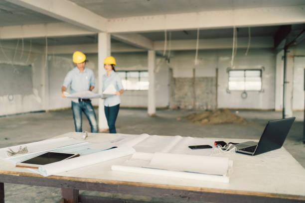 Blueprint, laptop computer, and industrial tools at construction site with two engineers or architects working together in blur background. Industry, building architecture, or teamwork concept