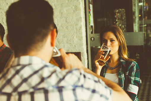 Young, beautiful, attractive and modern couple are in bar. They are sitting and drinking beer. Both are dressed casual, they are wearing check shirts. He have short black hair, while she is brunette and have long hair. Camera is behind his back. Behind couple is a group of people who also drink beer and have fun. All these people are in modern bar and have great time. Happiness and enjoyment is all around this space.