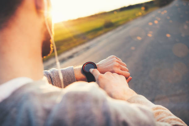 joven atleta corredor en auriculares mira un reloj elegante moderno y cuenta calorías pasó después al aire libre en la formación al atardecer - sun watch fotografías e imágenes de stock