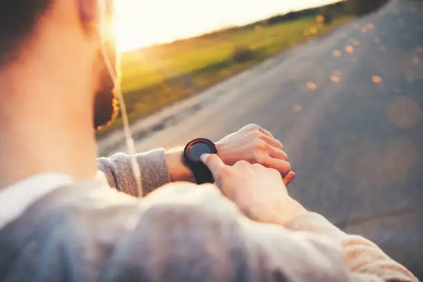 Photo of Young athlete runner in headphones looks at a modern smart clock and counts up spent calories after training outdoors at sunset