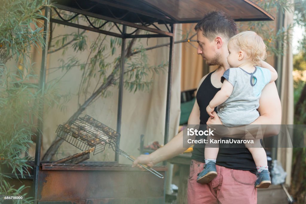 Father and son at barbecue grill in park Father and his baby boy son at barbecue grill. Father holds 1 year-old son on hands and preparing meat outdoor Barbecue - Meal Stock Photo