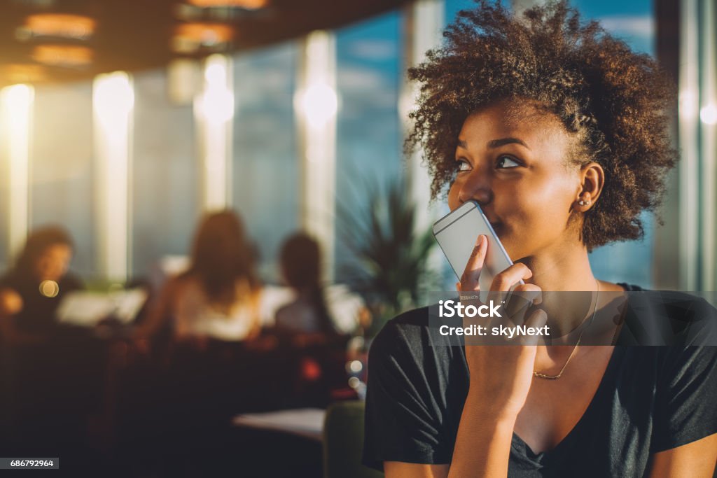 Curly black girl using her phone as voice recorder Young curly black female in luxury office space recording audio message via her smartphone, afro american teenage curly girl using her cell telephone as recorder to send voice message to her mother Mobile Phone Stock Photo