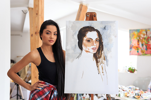 Female model posing next to her portrait in workshop. Looking at camera.