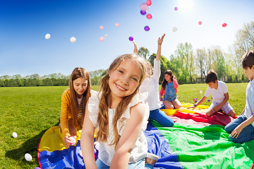 Close-up portrait of smiling fair-haired girl, sitting on rainbow parachute during the game with friends in the park