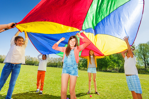 Joyful kids playing with rainbow parachute, standing under it's big canopy in the park