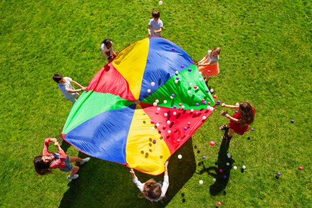 Happy kids waving rainbow parachute full of balls Top view picture of kids standing in a circle on the green lawn and holding rainbow parachute full of colorful balls summer camp stock pictures, royalty-free photos & images