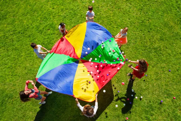 Top view picture of kids standing in a circle on the green lawn and holding rainbow parachute full of colorful balls