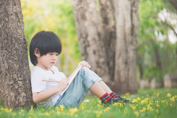 istock Little Asian boy sitting under the tree and drawing in notebook at park 686911754