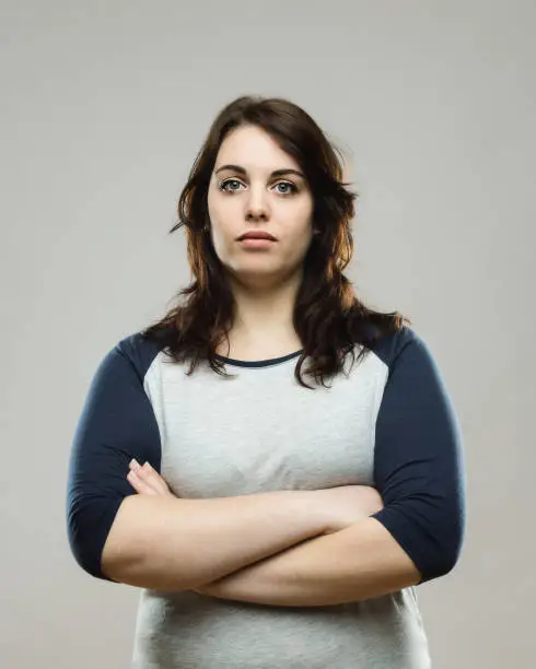 Portrait of confident young woman standing with her arms crossed against gray background. Vertical shot of healthy real woman in studio. Photography from a DSLR camera. Sharp focus on eyes.