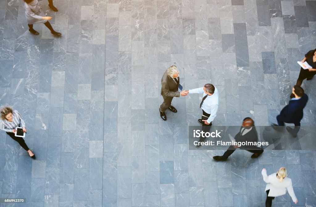 Adult office workers shaking hands in crowded hall From above two adult business men shaking hands met in office hall with workers walking around. High Angle View Stock Photo