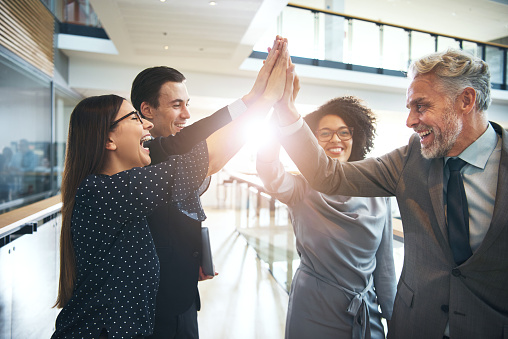 Smiling and laughing office working black and white men and women standing and giving high five to each other.