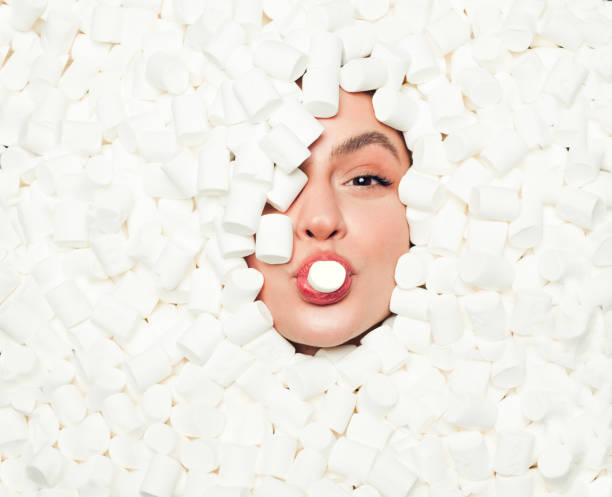 Woman biting a marshmallow From above female face lying in a pile of marshmallows and biting the one. candy in mouth stock pictures, royalty-free photos & images