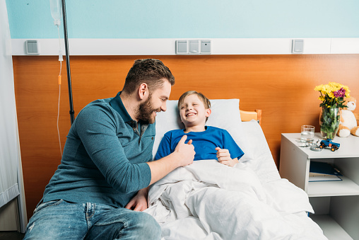 Smiling father playing with cute little son lying in hospital bed, dad and son in hospital