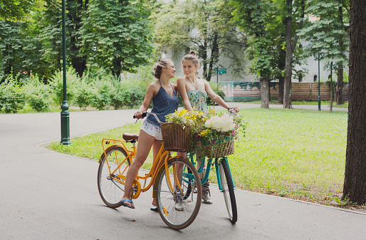 Happy boho girls gather wild flowers on bicycle ride. Beautiful female friends, youth and happiness, active summer leisure in park concept.