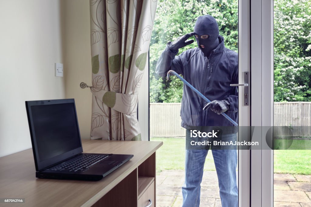 Thief looking through patio doors window at a laptop computer to steal Thief breaking into a house via a patio doors window with a crowbar to steal a laptop computer from an office desk Burglar Stock Photo