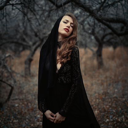 Beautiful girl in in black vintage dress with curly hair posing in the woods. Woman in retro dress lost in the forest. Worried sensual emotion . Retro fashion. Red lips.