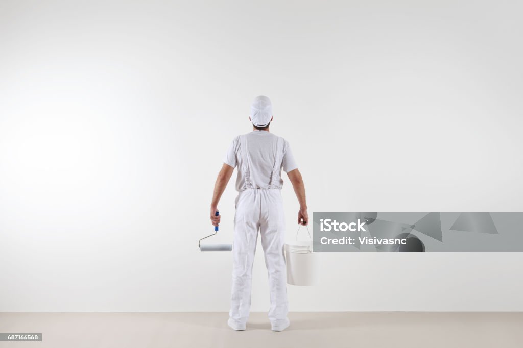 Vue arrière de l'homme de peintre regardant le mur blanc, avec le rouleau et le seau de peinture, d'isolement sur la pièce blanche - Photo de Peintre libre de droits