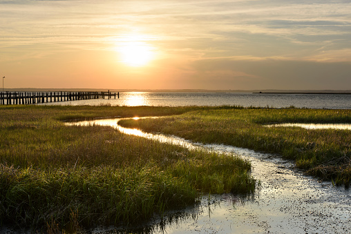 View of sunset over ocean inlet with green grasses and dock.