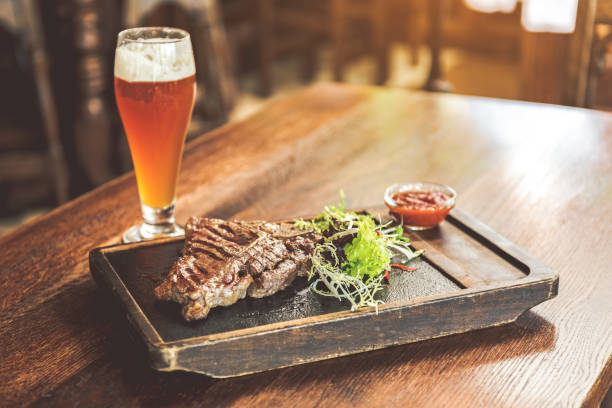 Spicy meat with light cold beer Delicious dish. Close up of fragrant grilled T-bone steak, mix salad and tomato sauce on cutting board with cool lager on wooden table. Pub in the background steak-and-ale stock pictures, royalty-free photos & images