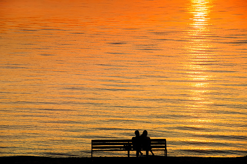 Silhouette of a couple on a bench at sunset against romantic orange colored sea. Summer leisure, vacation and romance theme