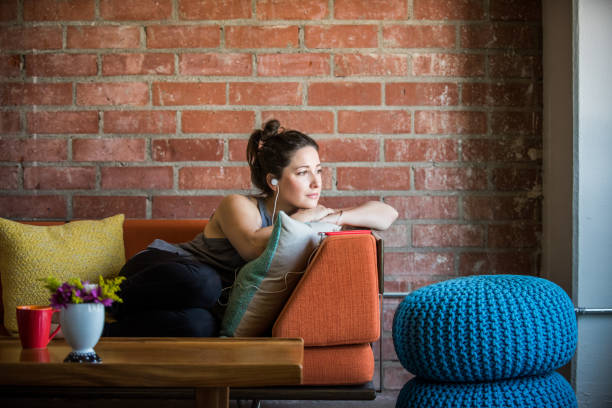 Young Woman Intently Listening to a Podcast Young caucasian woman intently listening to podcast, being reflective and introspective while lying on her side on her sofa in her loft apartment yoga pants photos stock pictures, royalty-free photos & images