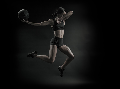 A desaturated image of an African Amercian women in mid-air with a basketball on a black background.  About to slam dunk it!