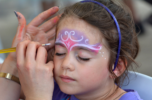 little girl (age 5-6) getting her face painted with a crown like a princes by face painting artist.