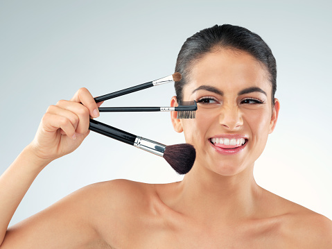 Studio shot of a beautiful young woman holding makeup brushes against a gray background
