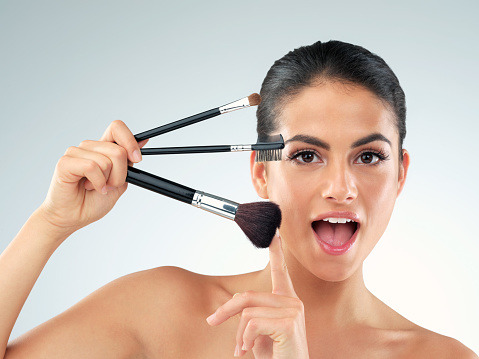 Studio shot of a beautiful young woman holding makeup brushes against a gray background