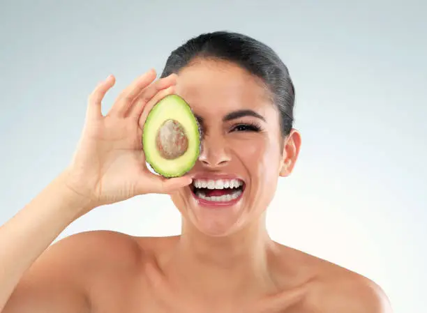Studio portrait of a beautiful young woman covering her eye with an avocado against a gray background