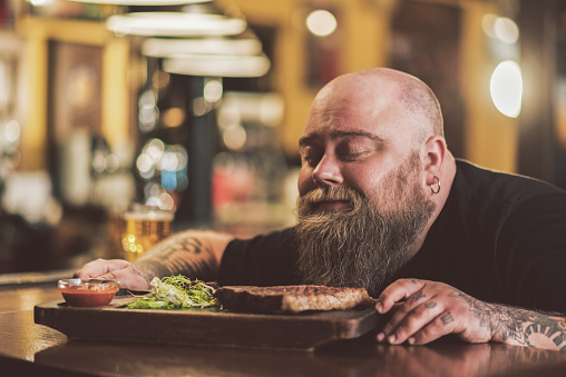 Divine aroma. face of pleasant bearded man sitting at bar counter and sniffing aromatic steak. He enjoying smell with closed eyes