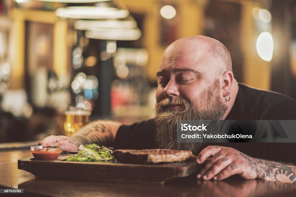 Obese male savoring grilled meat in pub Divine aroma. face of pleasant bearded man sitting at bar counter and sniffing aromatic steak. He enjoying smell with closed eyes Eating Stock Photo