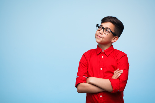 Adorable young man standing with arms crossed and daydreaming on the blue background.