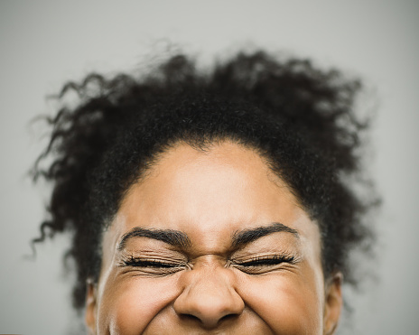 Close-up portrait of a real happy young afro american woman with beautiful toothy smile and closed eyes. Cheerful female is against gray background. Horizontal studio photography from a DSLR camera. Sharp focus on eyes.